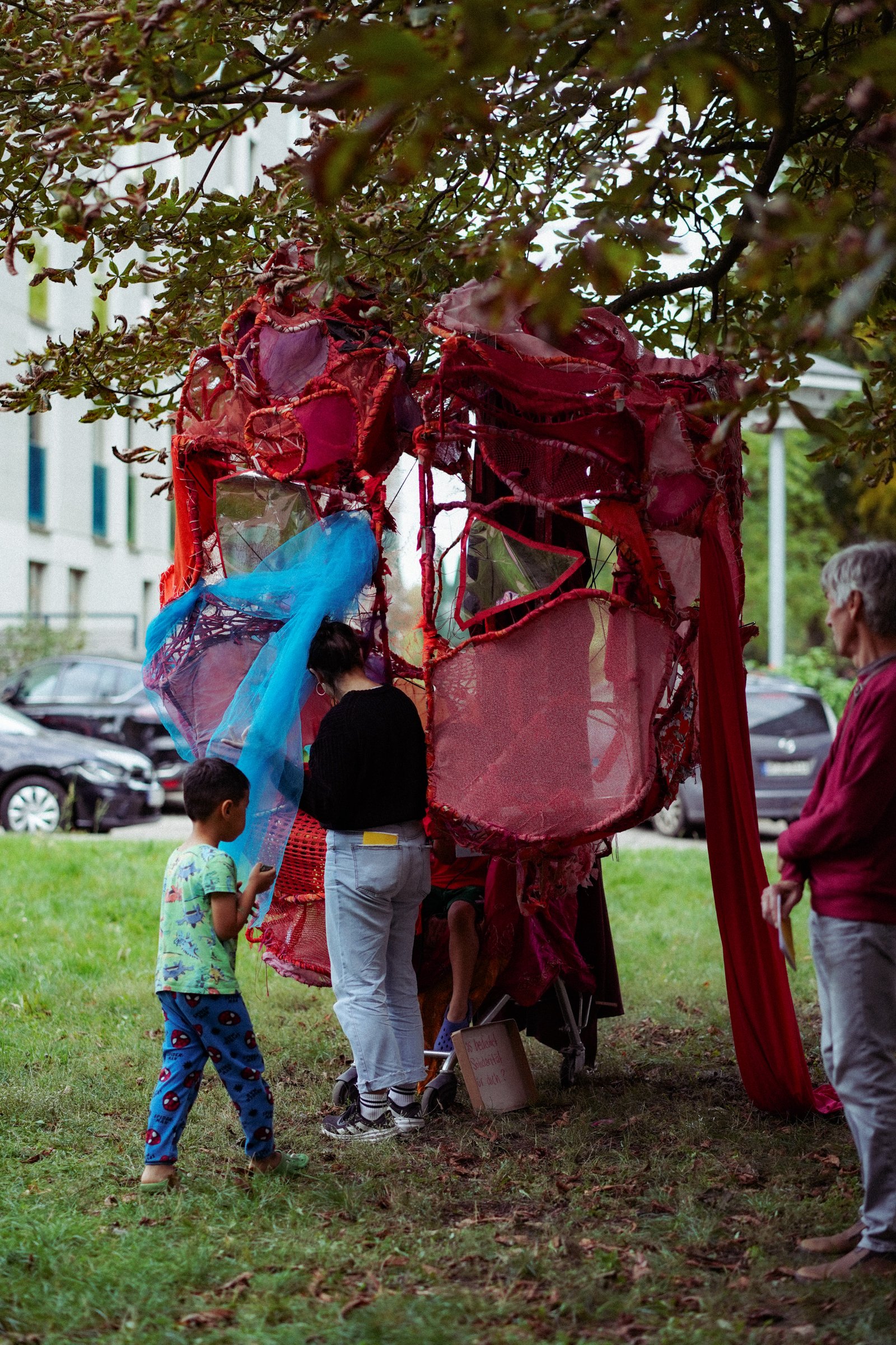 Eine große rote Stoffinstallation unter einem Baum, Kinder und Erwachsene schauen hinein.