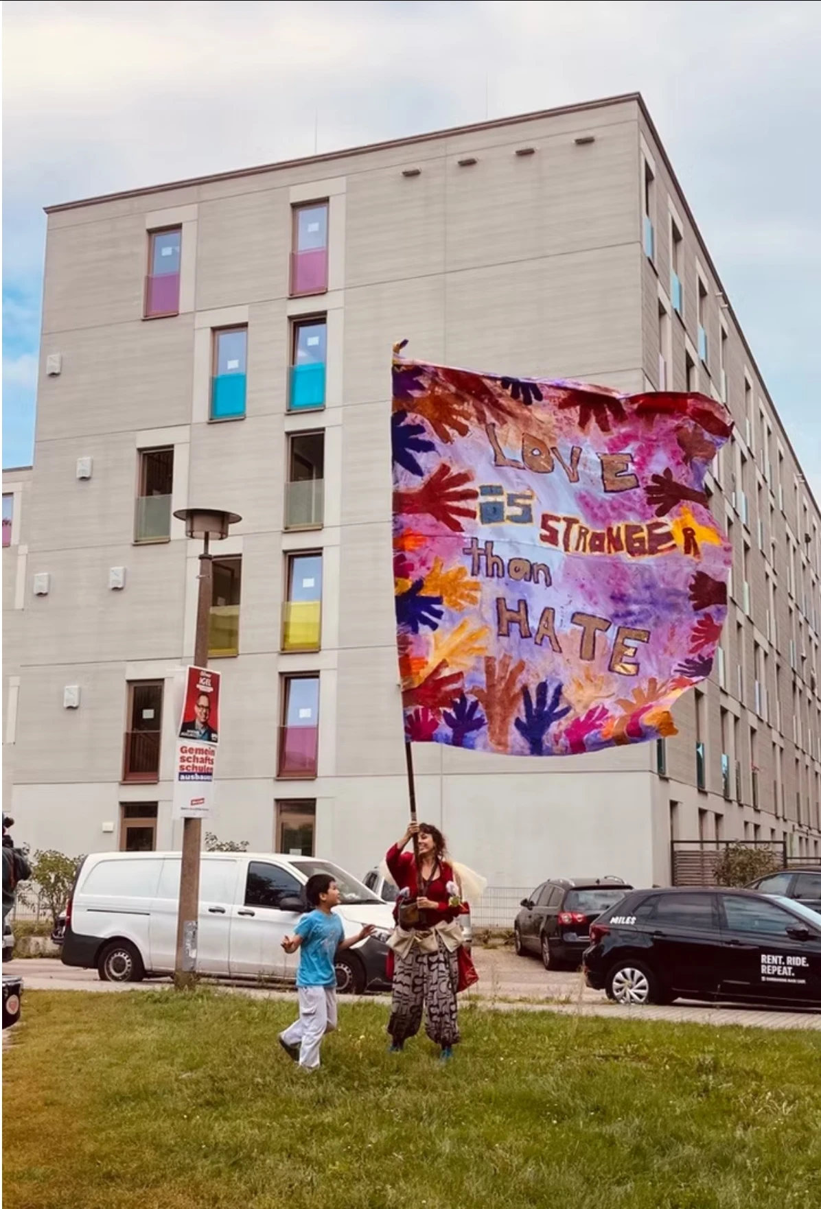 Vor einem Wohnhaus schwenkt eine Person eine riesige bemalte Flagge mit der Aufschrift „Love is stronger than hate“, ein Kind läuft daneben.