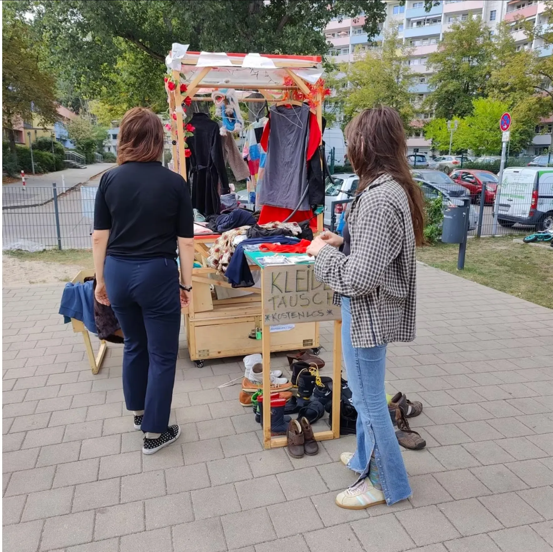 Zwei Personen an einem mobilen Holzstand mit Kleidung und Schuhen, auf dem Schild steht „Kleidertausch – kostenlos“.